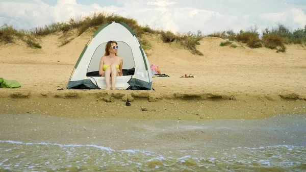 Ένα κορίτσι με μαγιό κοιτάζει έξω από μια σκηνή camping σε μια αμμώδη παραλία με φόντο τη θάλασσα και τους αμμόλοφους το καλοκαίρι. έννοια της ενότητας με τη φύση — Φωτογραφία Αρχείου