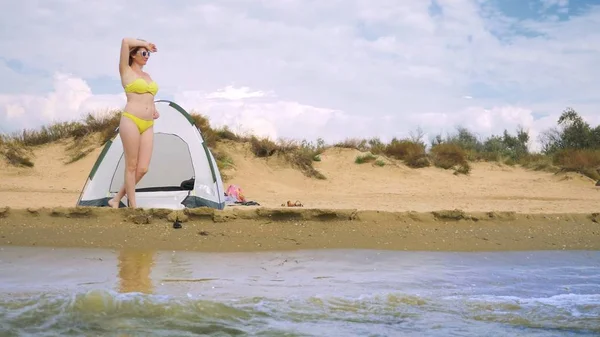 a girl in a swimsuit is resting next to a camping tent on a sandy beach against the background of the sea and dunes in the summer. concept of unity with nature