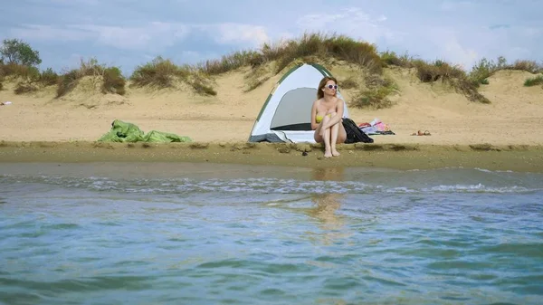 Ένα κορίτσι με μαγιό αναπαύεται δίπλα σε μια σκηνή κατασκήνωσης σε μια αμμώδη παραλία με φόντο τη θάλασσα και τους αμμόλοφους το καλοκαίρι. έννοια της ενότητας με τη φύση — Φωτογραφία Αρχείου