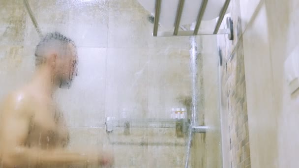 Młody przystojny człowiek chusteczki ręcznik stojąc pod prysznicem. widok przez szklankę. — Wideo stockowe
