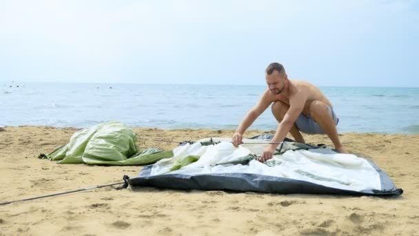 Τύπος με σορτς δημιουργεί μια σκηνή για κάμπινγκ σε μια αμμώδη παραλία με φόντο τη θάλασσα το καλοκαίρι — Αρχείο Βίντεο