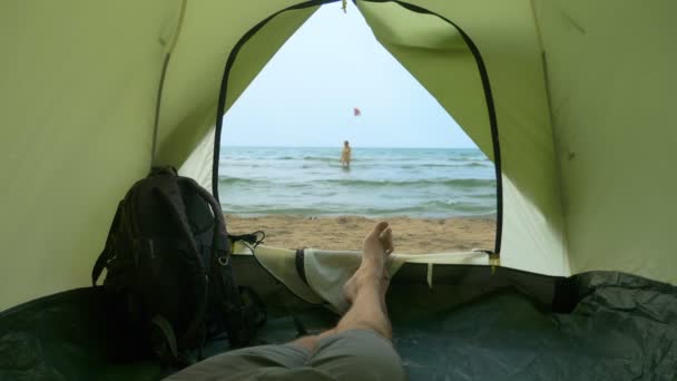 Konzept des Zeltens am Meer. ein Mann beobachtet aus einem Zelt seine Freundin, die im Meer planscht — Stockvideo