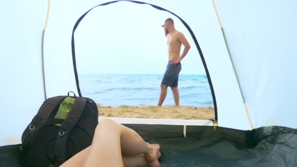 Concepto de camping junto al mar. vista desde la tienda, la chica observa a su novio salpicando en el mar, acostado en la tienda — Vídeo de stock