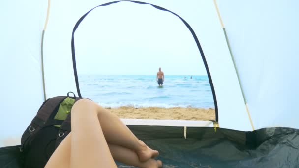 海辺でのキャンプコンセプト。テントから見ると、女の子は彼氏が海に飛び散り、テントの中に横たわっているのを見ている — ストック動画