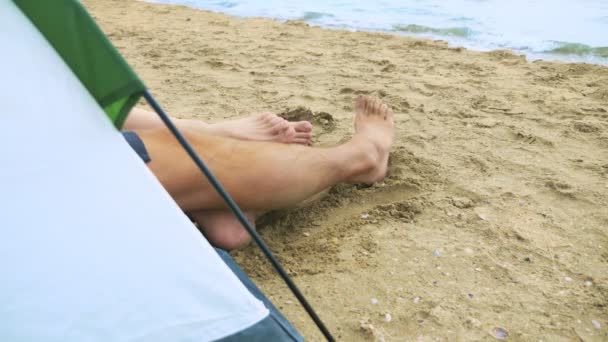 Camping konceptet vid havet. benen på en man och kvinna är synliga från ett tält på sanden mot bakgrunden av havet. — Stockvideo