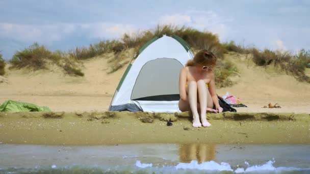 En flicka i en baddräkt vilar bredvid en camping tält på en sandstrand mot bakgrund av havet och sanddyner på sommaren. begreppet enhet med naturen — Stockvideo
