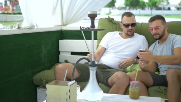 Una pareja gay fuma una cachimba y mira la aplicación por teléfono, sentada en el cenador en la calle en un día soleado de verano . — Vídeos de Stock