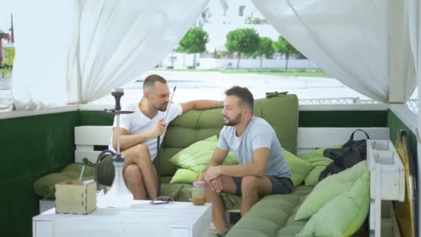Schwules Paar raucht Wasserpfeife und sitzt an einem sonnigen Sommertag im Pavillon auf der Straße. — Stockvideo