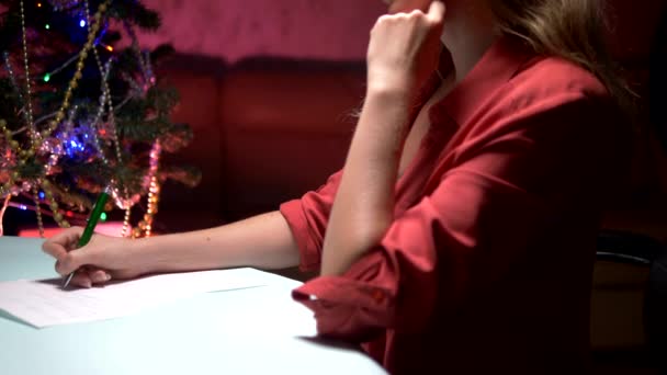 女性は装飾されたクリスマスツリーの近くのテーブルに座って、サンタクロースにクリスマスレターを書きます — ストック動画