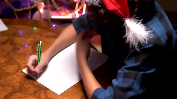 Tiener jongen zit aan een tafel in de buurt van een versierde kerstboom en schrijft een Nieuwjaar brief aan de Kerstman — Stockvideo