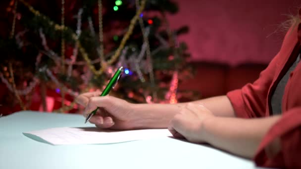 Mulher senta-se a uma mesa perto de uma árvore de Natal decorada e escreve uma carta de Natal para Papai Noel — Vídeo de Stock