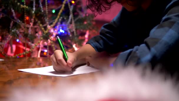 Adolescente se sienta en una mesa cerca de un árbol de Navidad decorado y escribe una carta de Año Nuevo a Santa Claus — Vídeo de stock