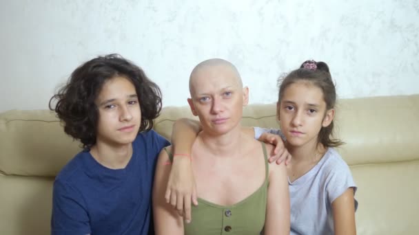 Лысая измученная мать после химиотерапии рака обнимает своих подростков, сидящих на диване — стоковое видео