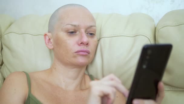 Mujer calva utiliza un teléfono inteligente mientras está sentado en un sofá — Vídeo de stock