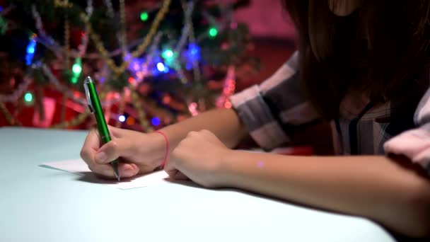 Mulher senta-se a uma mesa perto de uma árvore de Natal decorada e escreve uma carta de Natal para Papai Noel — Vídeo de Stock