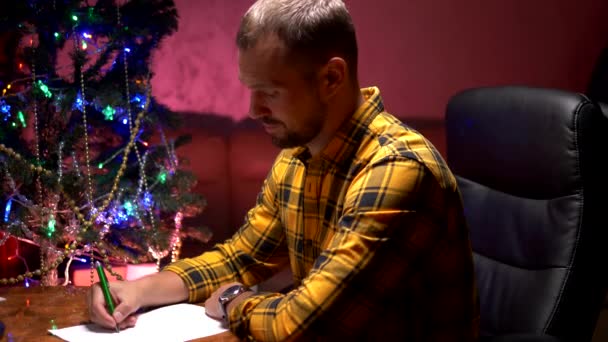 Чоловік сидить за столом біля прикрашену ялинку і пише новий рік листа Санта-Клауса — стокове відео