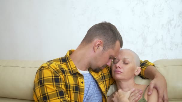 Лысая женщина и мужчина обнимаются, сидя на диване. Любовь пара преодолевает ее онкологию вместе — стоковое видео
