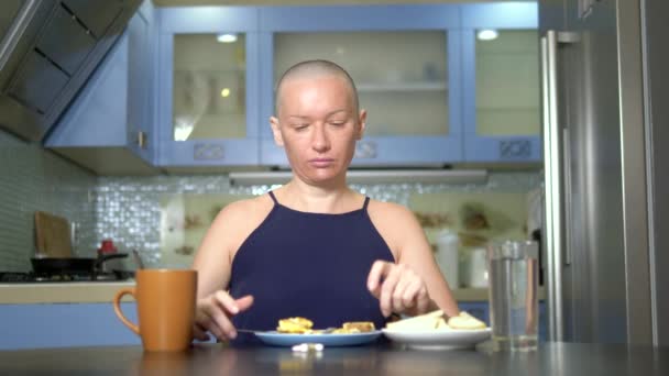 Лысая грустная женщина сидит за столом на кухне с едой и таблетками, неохотно завтракает, чувствует тошноту — стоковое видео