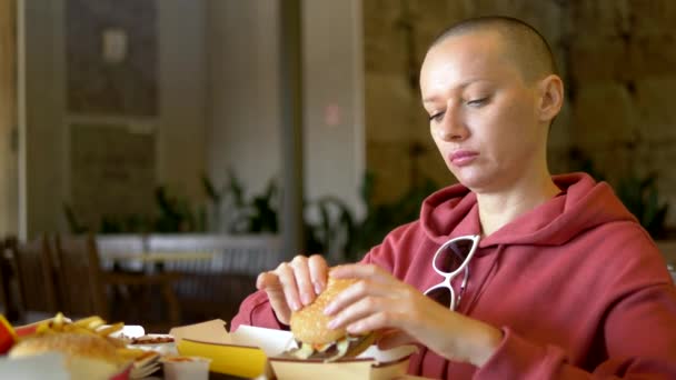 Hambrienta mujer calva comiendo una sabrosa hamburguesa en un restaurante de comida rápida . — Vídeos de Stock