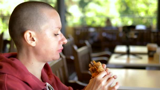 Una ragazza pelata in un bar che mangia un hamburger. primo piano. guardando la macchina fotografica — Video Stock