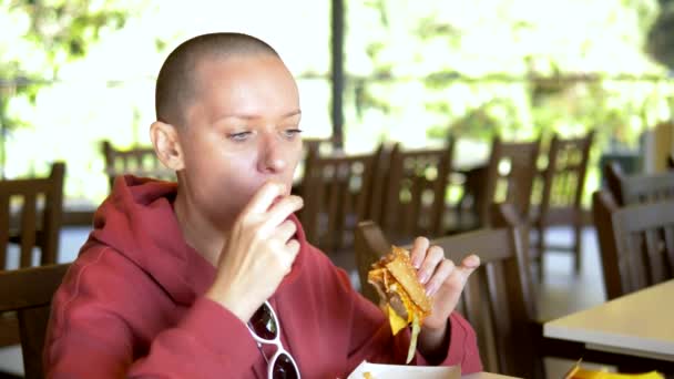Φαλακρή κοπέλα σε ένα καφέ που τρώει χάμπουργκερ. Κοντινό πλάνο. Κοιτάζοντας την κάμερα — Αρχείο Βίντεο