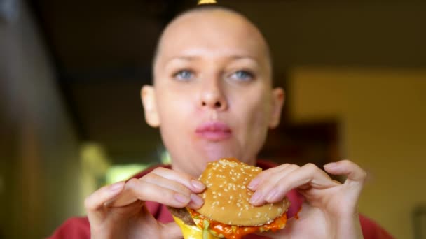Una ragazza pelata in un bar che mangia un hamburger. primo piano. guardando la macchina fotografica — Video Stock