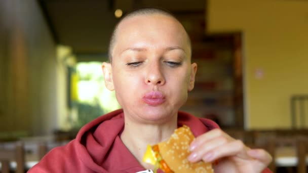 Лысая девушка в кафе ест гамбургер. крупным планом. глядя в камеру — стоковое видео