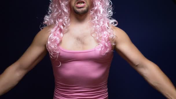 Verrückter gutaussehender junger Kerl mit lockiger Perücke und rosa T-Shirt vor dunklem Hintergrund tanzt lustig, zeigt seine Muskeln — Stockvideo