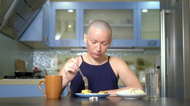 bald sad woman sits at a table in the kitchen with food and pills, reluctantly eats breakfast, feeling nauseous clipart