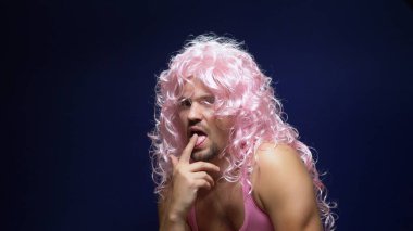Kıvırcık peruklu ve pembe tişörtlü çılgın yakışıklı adam arka planda dans ediyor, kaslarını gösteriyor.