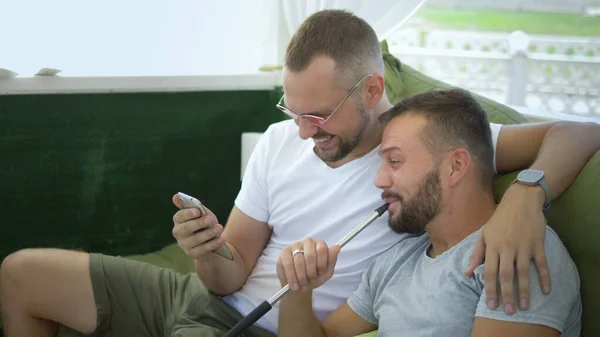 Um casal gay fuma um narguilé e observa o aplicativo no telefone, sentado no gazebo na rua em um dia ensolarado de verão . — Fotografia de Stock