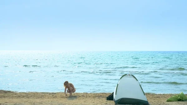 Campingkonzept am Meer. Mädchen sammelt Muscheln am Strand in der Nähe des Zeltes auf dem Meeresgrund. — Stockfoto