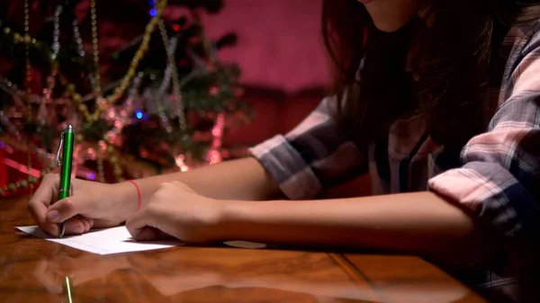 Menina adolescente senta-se em uma mesa perto de uma árvore de Natal decorada e escreve uma carta de Natal para Papai Noel — Fotografia de Stock