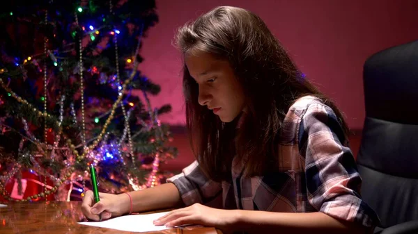 십대 소녀는 장식 된 크리스마스 트리 근처 테이블에 앉아 산타 클로스에게 크리스마스 편지를 씁니다. — 스톡 사진