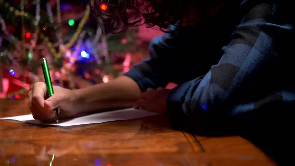 Adolescente menino senta-se a uma mesa perto de uma árvore de Natal decorada e escreve uma carta de Ano Novo para Papai Noel — Fotografia de Stock