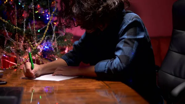 Підліток хлопчик сидить за столом біля прикрашену ялинку і пише новий рік лист Санта-Клауса — стокове фото