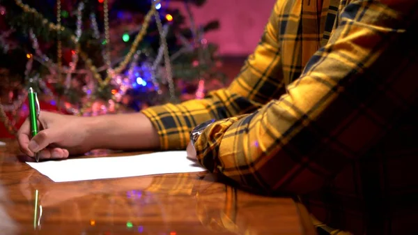 Um homem senta-se em uma mesa perto de uma árvore de Natal decorada e escreve uma carta de Ano Novo para Papai Noel — Fotografia de Stock