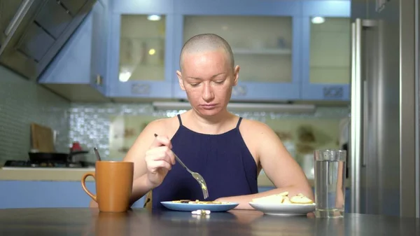 bald sad woman sits at a table in the kitchen with food and pills, reluctantly eats breakfast, feeling nauseous