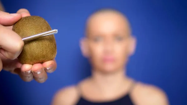 Atractiva mujer calva afeita un kiwi con una navaja peligrosa. concepto de humor, aventuras de gente extraña . — Foto de Stock