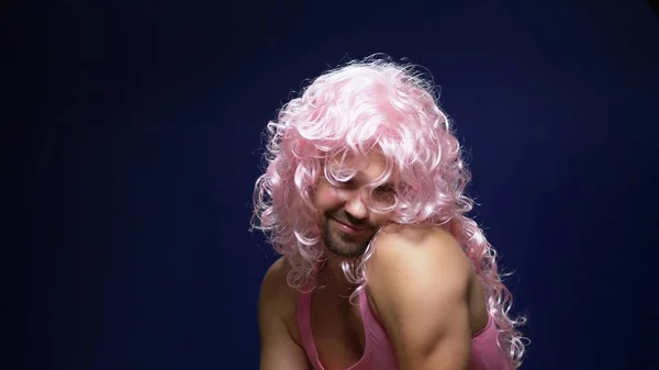 Louco bonito jovem cara em uma peruca encaracolado e uma camiseta rosa contra um fundo escuro está dançando engraçado, mostra seus músculos — Fotografia de Stock