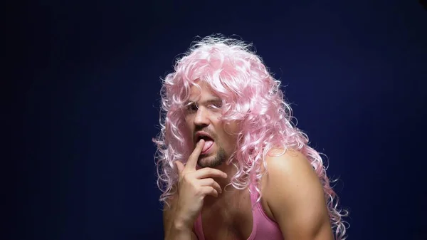 crazy handsome young guy in a curly wig and a pink t-shirt against a dark background is dancing funny, shows his muscles