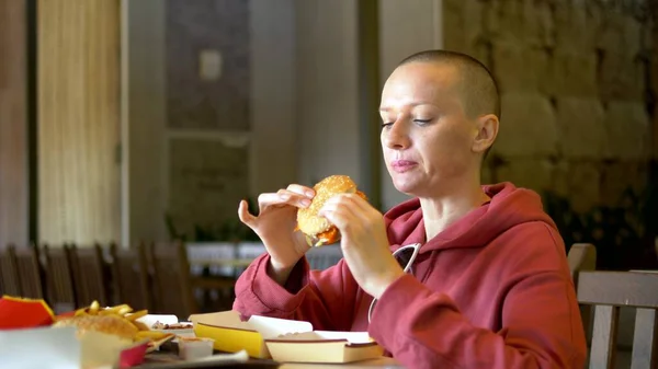 Nälkäinen kalju nainen syö maukasta hampurilaista pikaruokaravintolassa . — kuvapankkivalokuva