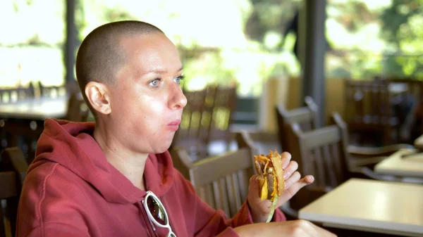 Hambrienta mujer calva comiendo una sabrosa hamburguesa en un restaurante de comida rápida . — Foto de Stock