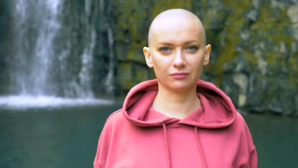 Mulher careca bonita olhando para a câmera, de pé no fundo de uma cachoeira. conceito de liberdade, vitória, realização de metas. espaço de cópia — Vídeo de Stock