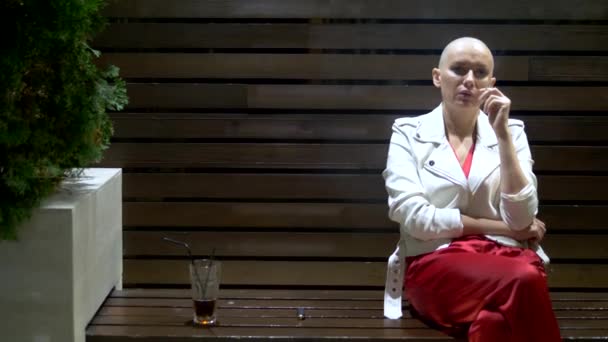 Chica calva en una chaqueta blanca de cuero fuma y bebe en la terraza en un club nocturno — Vídeos de Stock