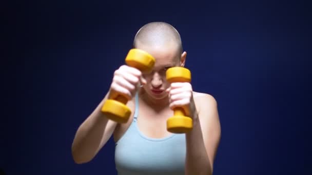 Bald woman boxing to the camera with dumbbells in hands on a dark background. — Stock Video