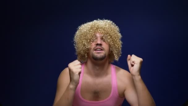 Louco bonito jovem cara em uma peruca encaracolado e uma camiseta rosa contra um fundo escuro está dançando engraçado, mostra seus músculos — Vídeo de Stock