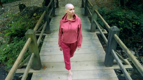 Mujer calva camina sobre un puente de madera en el bosque. El concepto de transición en la vida . — Vídeos de Stock