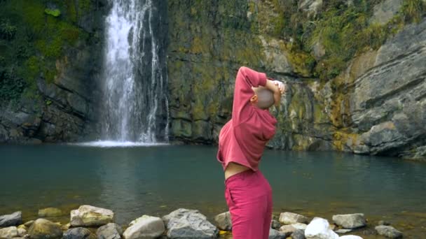 Bald young woman standing in front of a waterfall with hands up. concept of freedom, victory, goal achievement. copy space — Stock Video