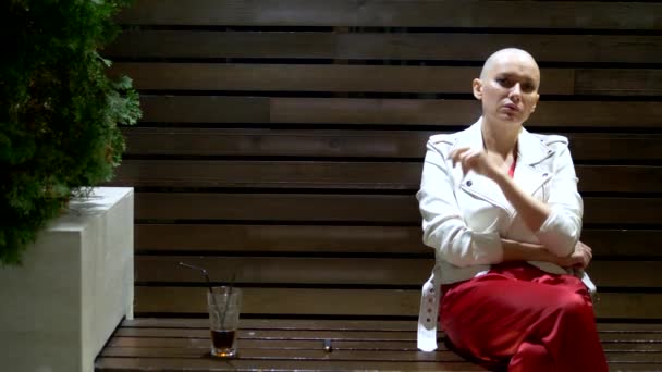 Chica calva en una chaqueta blanca de cuero fuma y bebe en la terraza en un club nocturno — Vídeo de stock